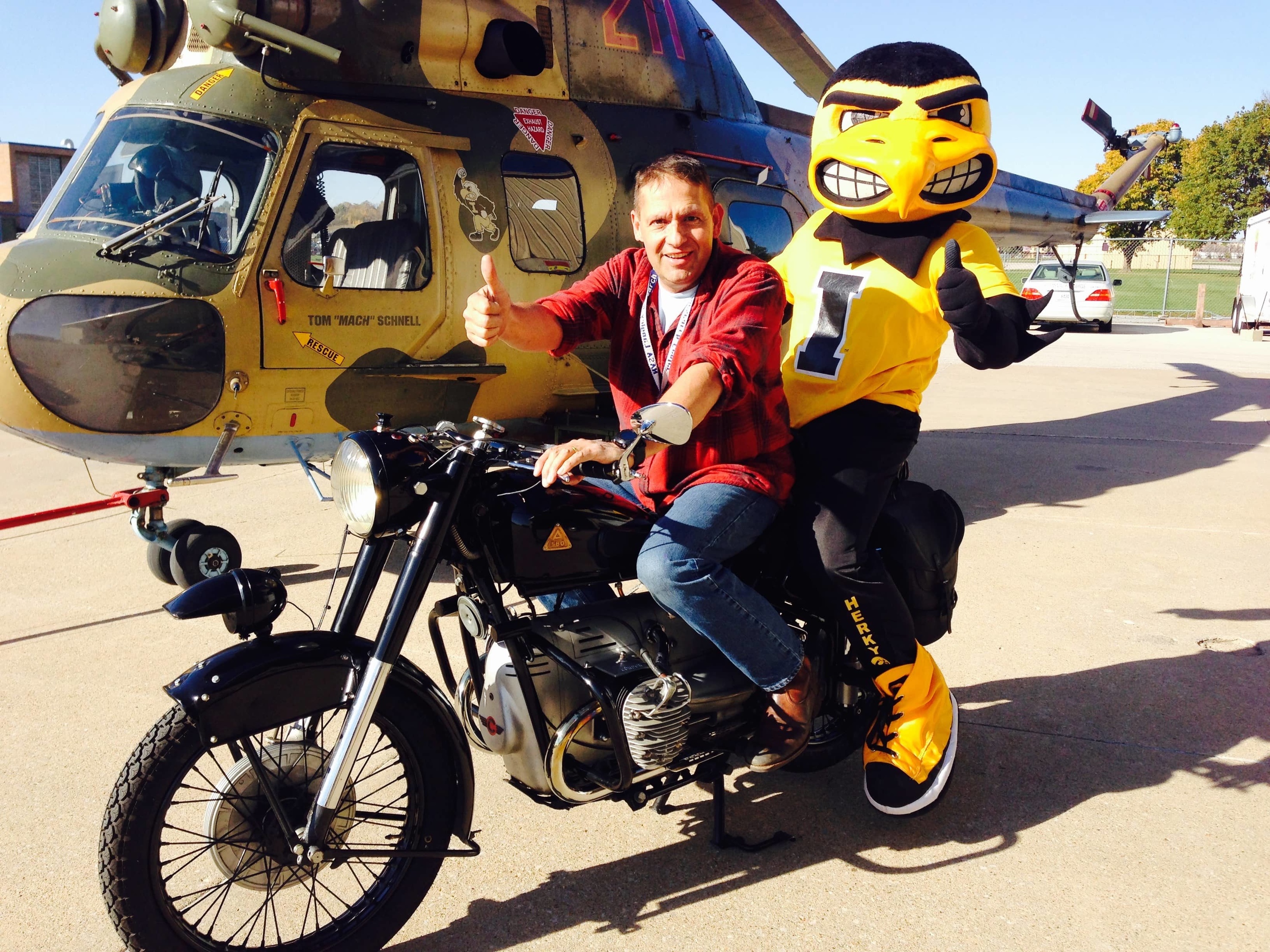 Tom Schnell and Herky on a motorcycle with a helicopter in the background